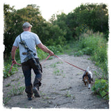 A man wearing a gray carbon hound t-shirt and dark pants walks on a dirt path with a dog on a leash, carrying a rifle on his back. The carbon hound t-shirt says Raise Your Spirits printed on the back.
