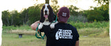desktop sized image for carbon hound about us page showing a man in a black Carbon Hound t-shirt that says Raise Your Spirits, holding a black, white and brown basset hound with a green leash, standing in a field