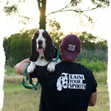 mobile image for carbon hound about us page showing a man in a black Carbon Hound t-shirt that says Raise Your Spirits, holding a black, white and brown basset hound with a green leash, standing in a field